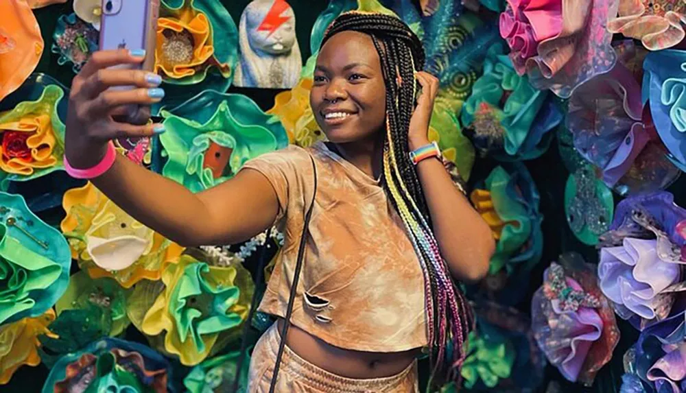 A person with braided hair and a rainbow wristband is smiling while taking a selfie against a vibrant backdrop of colorful abstract shapes