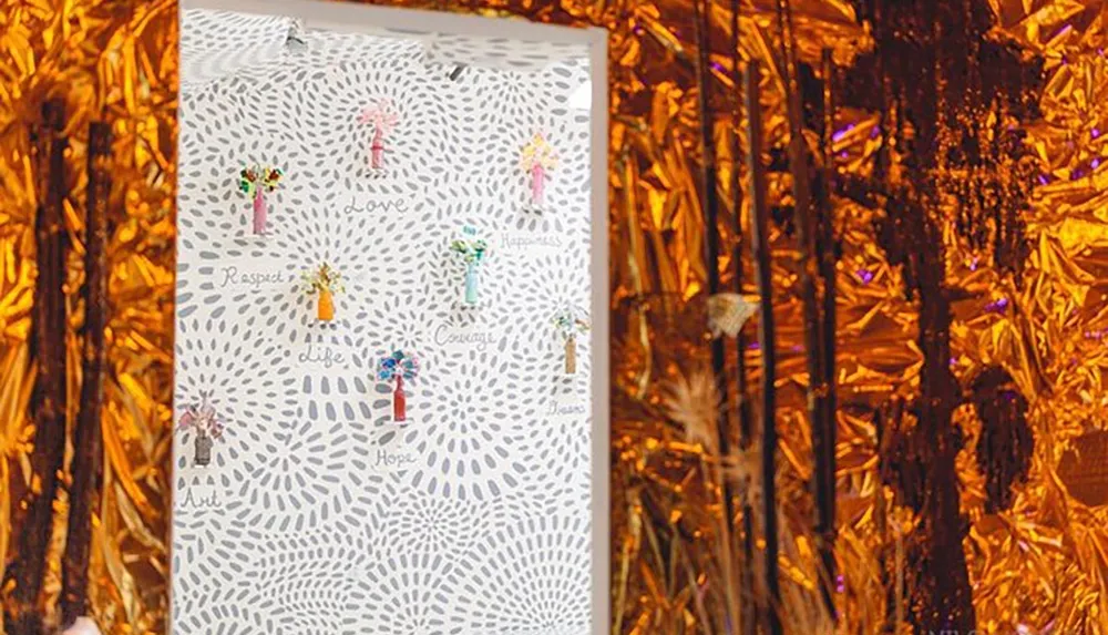 The image shows an artwork featuring colorful abstract human-like figures and words such as love hope and dreams on a patterned background juxtaposed with a reflective metallic gold curtain on the right