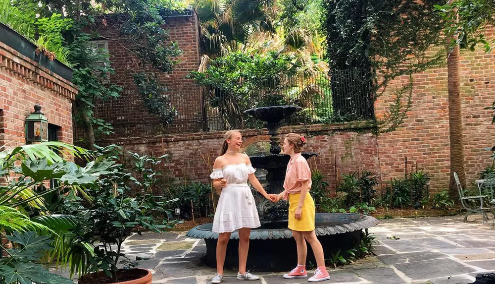 Two people are holding hands and smiling at each other in a lush courtyard with a fountain surrounded by brick walls and tropical plants