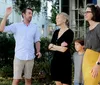A group of four people possibly a family stand outdoors with a man gesticulating as if explaining something a smiling woman looking at him another woman watching something in the distance and a child standing between the adults