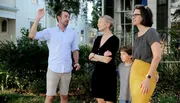 A group of four people, possibly a family, stand outdoors with a man gesticulating as if explaining something, a smiling woman looking at him, another woman watching something in the distance, and a child standing between the adults.