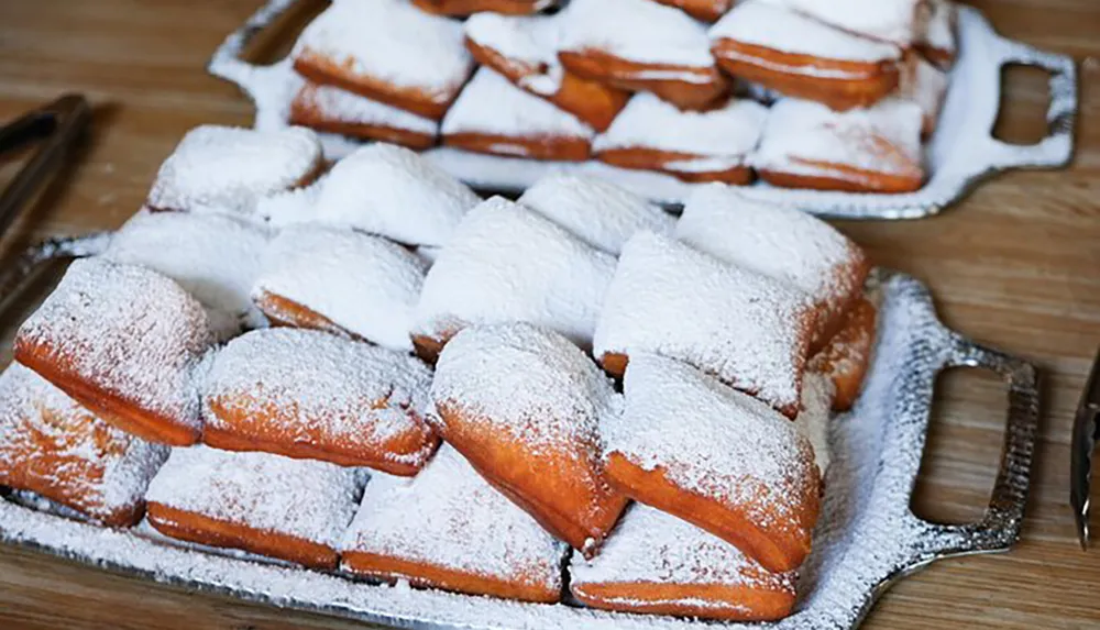 A batch of freshly made beignets is generously dusted with powdered sugar and presented on a metal tray