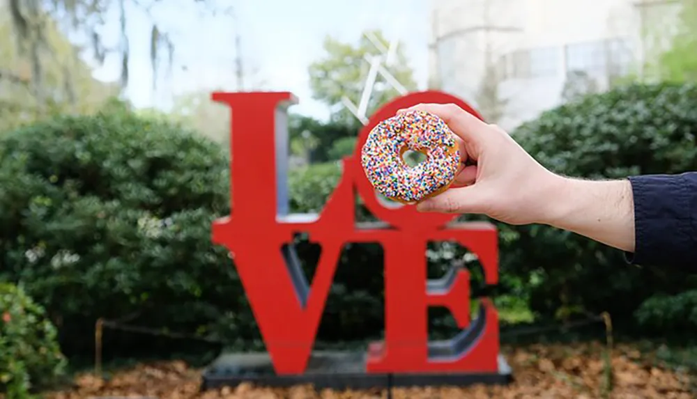 A persons hand is holding a sprinkle-covered donut in front of a large LOVE sign creatively positioning the donut to represent the letter O