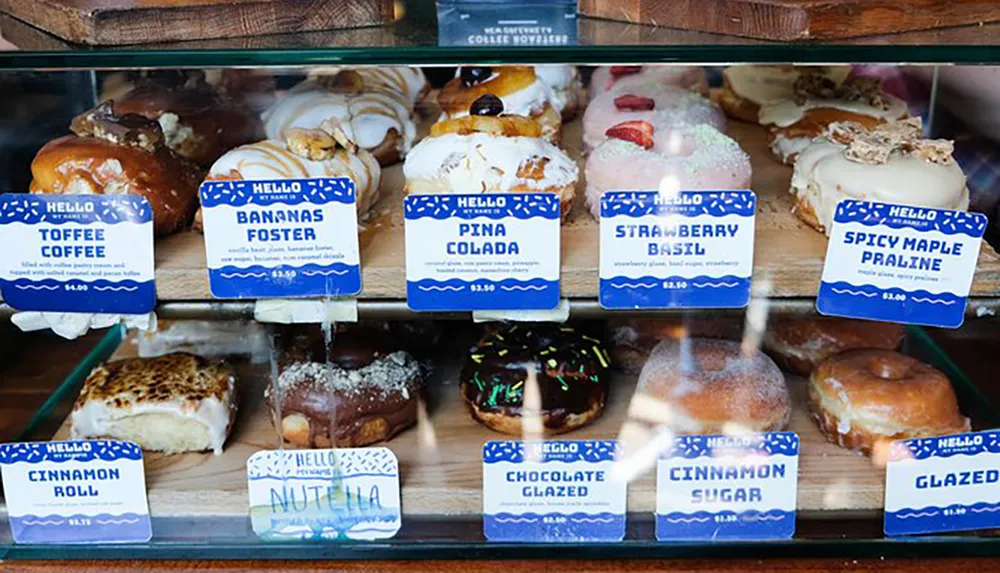 A variety of gourmet doughnuts with unique flavors like Bananas Foster Pina Colada and Strawberry Basil are displayed in a bakery case with labeled prices