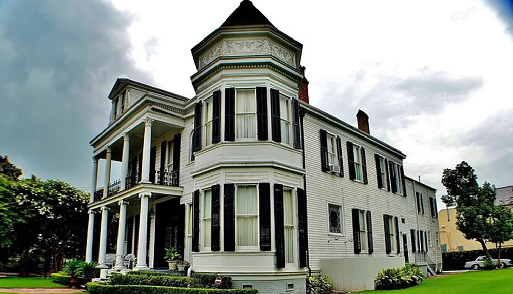 The image shows a large two-story white house with a classic Victorian architecture featuring a prominent bay window on the front corner and a spacious porch with columns