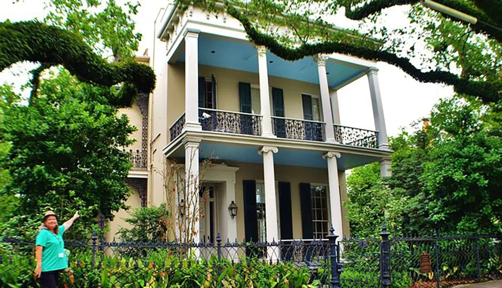 A person is smiling and pointing at a two-story house with blue walls a balcony with iron railings and surrounded by lush vegetation including a large tree with spreading branches to the left