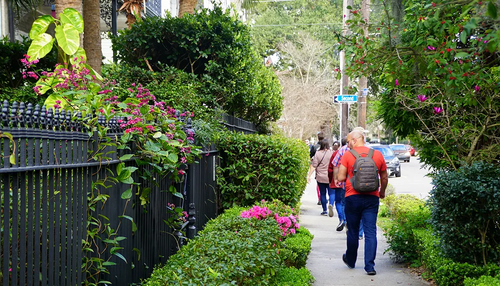 People walk down a lush plant-lined sidewalk next to a black metal fence under a sign for Second Street