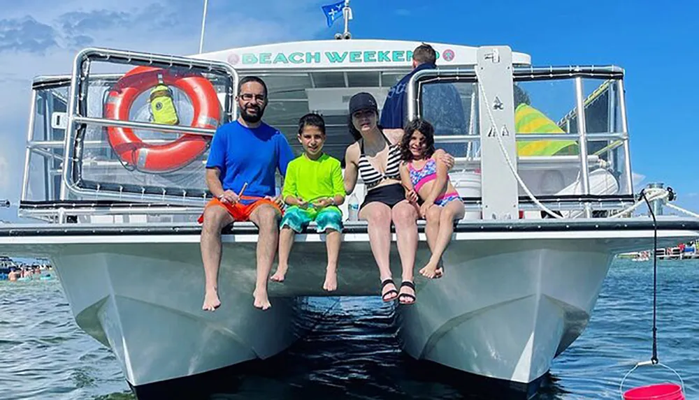 A family is sitting at the bow of a catamaran style boat named BEACH WEEKER smiling and posing for a photo on a sunny day