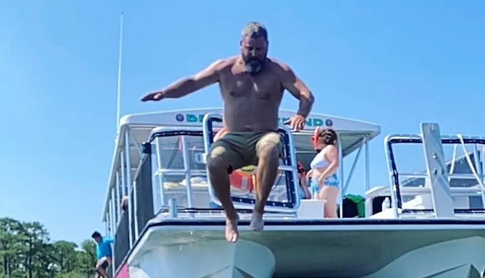 A shirtless man with shorts is mid-jump off the top of a boat under a clear blue sky while another person watches from the boat deck