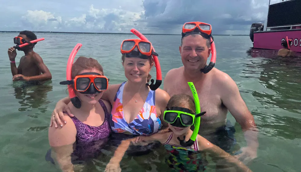 A family wearing snorkeling gear is posing for a photo in shallow waters with a boat in the background