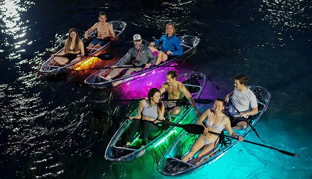 A group of people enjoy a nighttime kayaking excursion in clear kayaks illuminated with colorful underwater lights