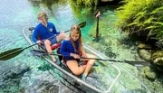 Two people are kayaking in clear water above an underwater scene of rocks and plants, with the kayak's clear bottom offering a view into the pristine aquatic environment.