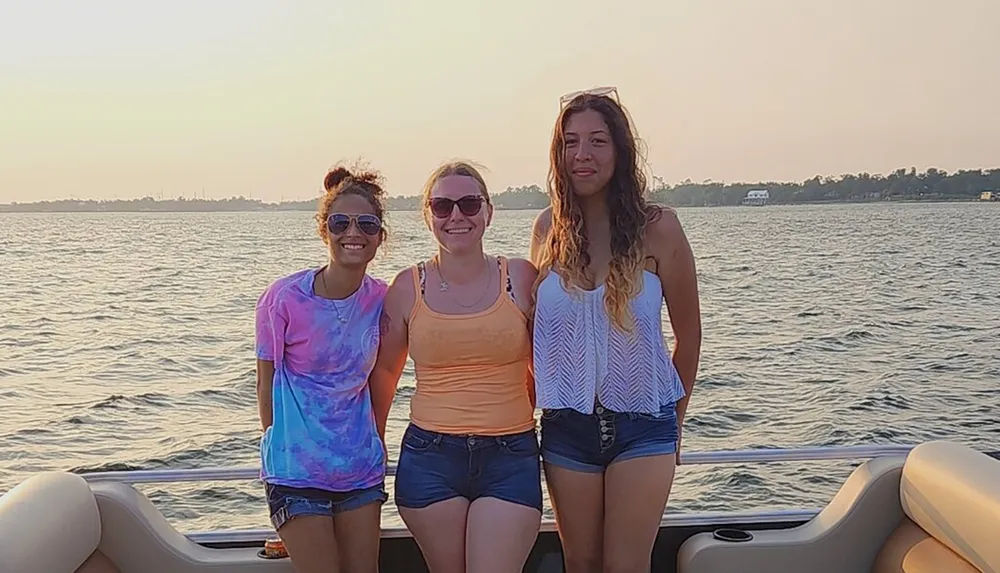 Three smiling women stand together on a boat enjoying the sunlight with a backdrop of calm waters