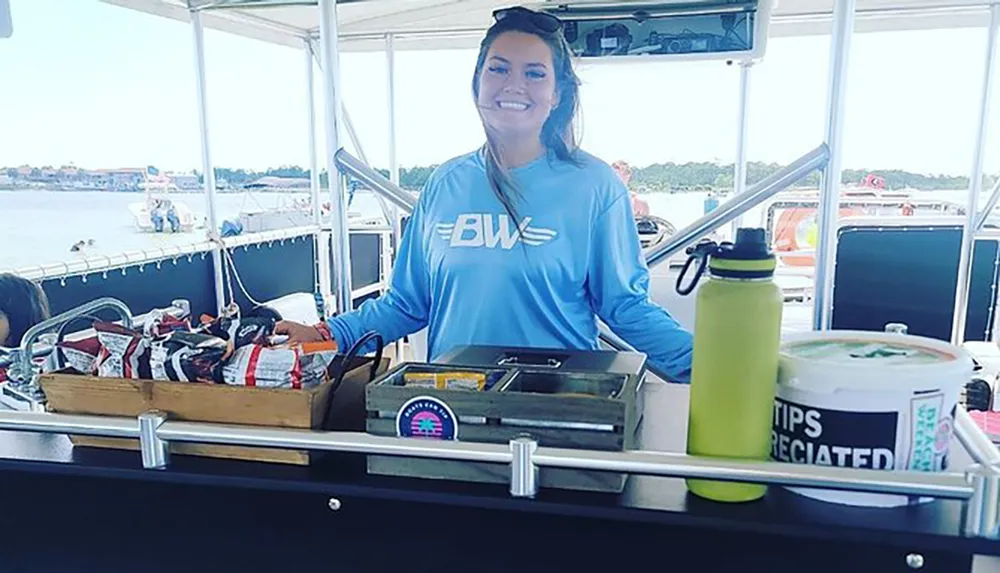 A smiling woman in a blue shirt stands behind a concession stand on a boat with snacks and a tip jar labeled TIPS APPRECIATED visible