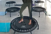 A person stands barefoot on a mini trampoline, with other trampolines visible in the background, suggesting this is a fitness or exercise setting.