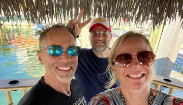 Three smiling people are posing for a selfie under a thatched structure with a man playfully giving a peace sign behind the head of another, reflecting a relaxed and cheerful atmosphere near a body of water with boats.