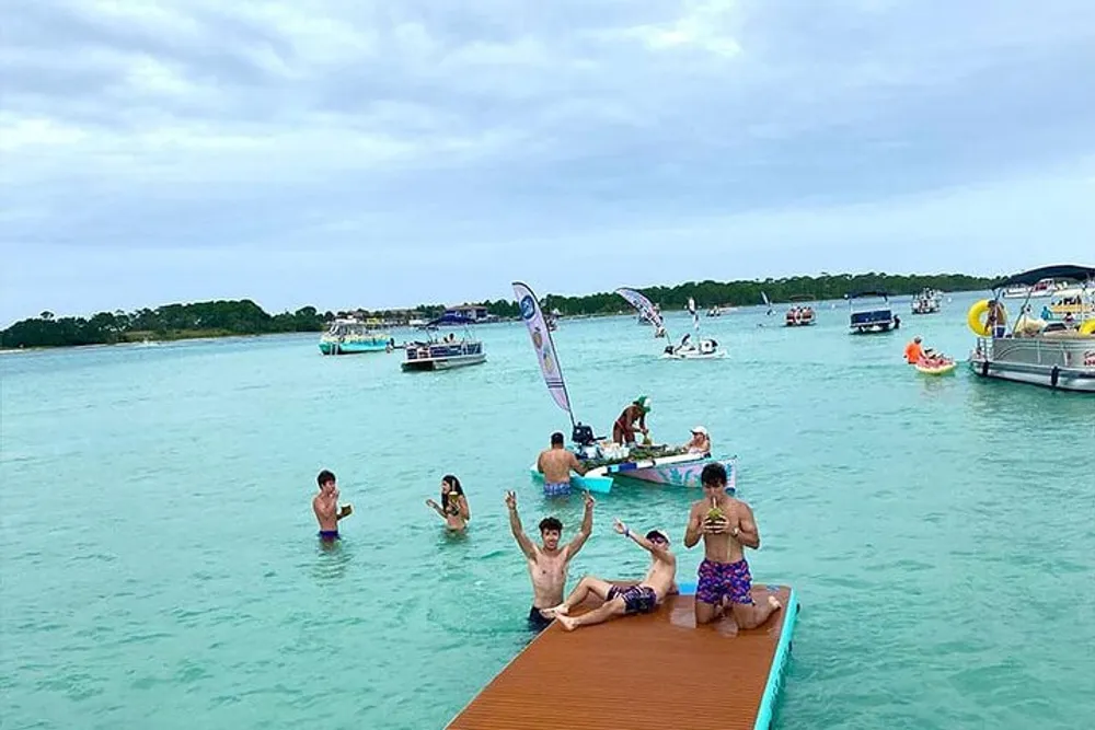 A group of people enjoy leisure time on a floating platform in crystal-clear waters surrounded by boats and overcast skies