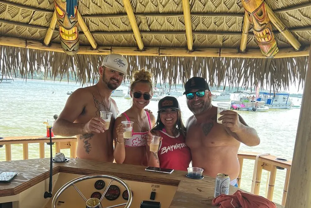 Four people are smiling and posing with drinks in hand at a tiki-style beach bar with a view of the water and boats in the background