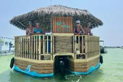 A group of people is enjoying a sunny day on a unique tiki bar-themed boat floating on calm water.