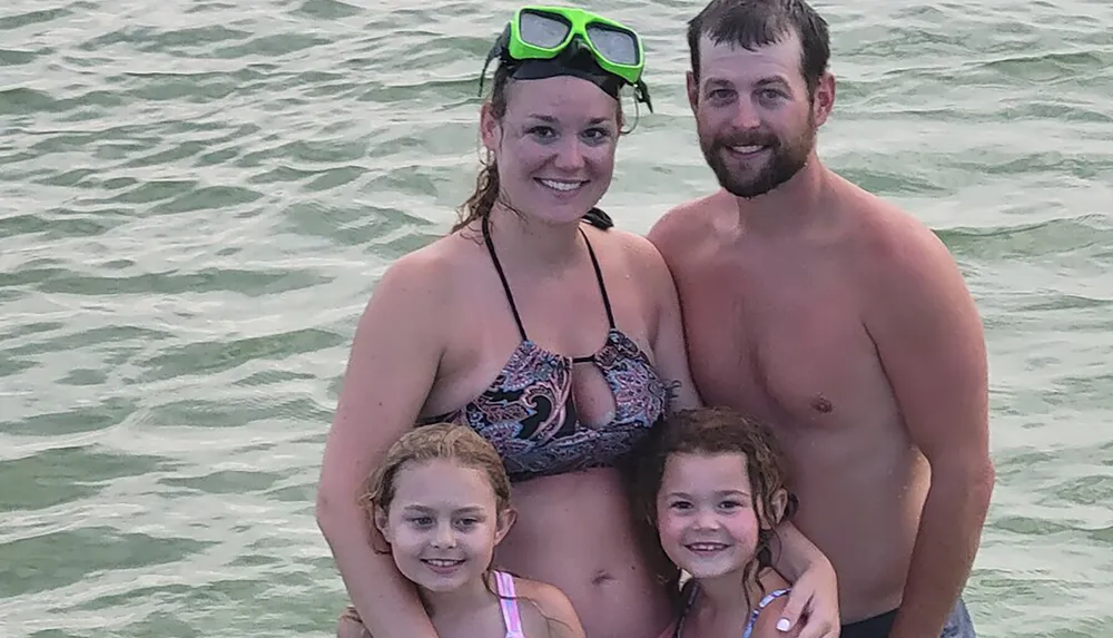 A family of four is posing for a photo in the water with the two adults standing behind two smiling children and one of the adults is wearing a pair of swim goggles on their head