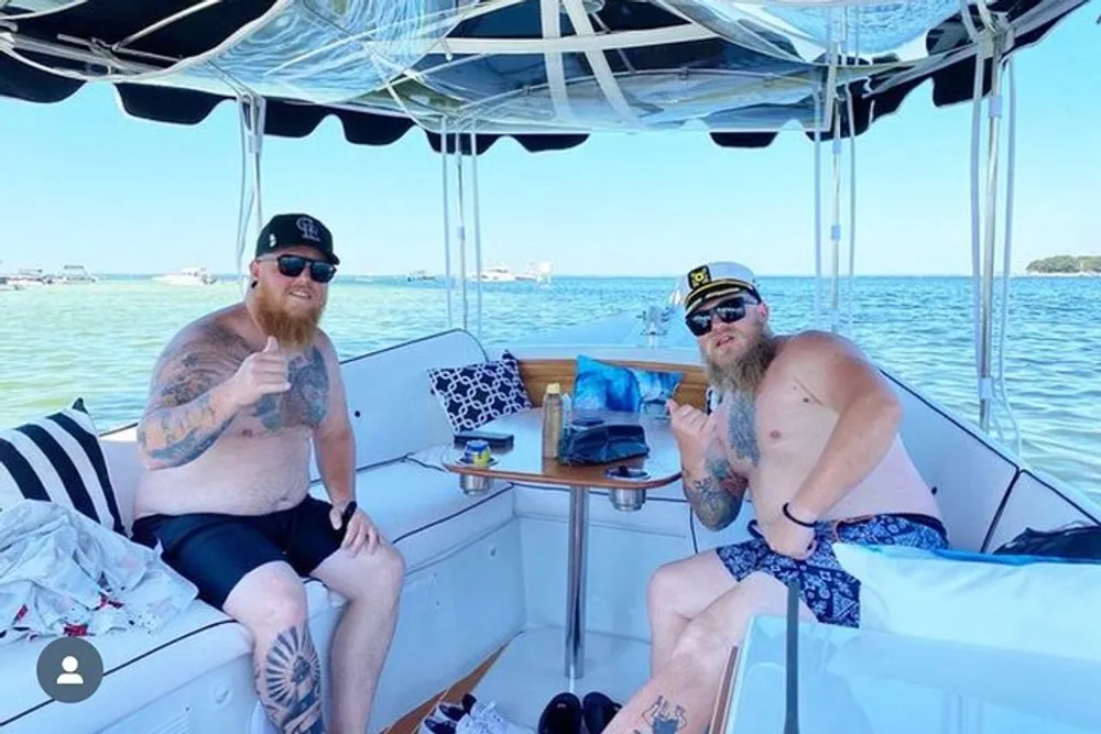 Two individuals with tattoos are sitting on a boat giving thumbs-up signs with clear skies and calm water in the background
