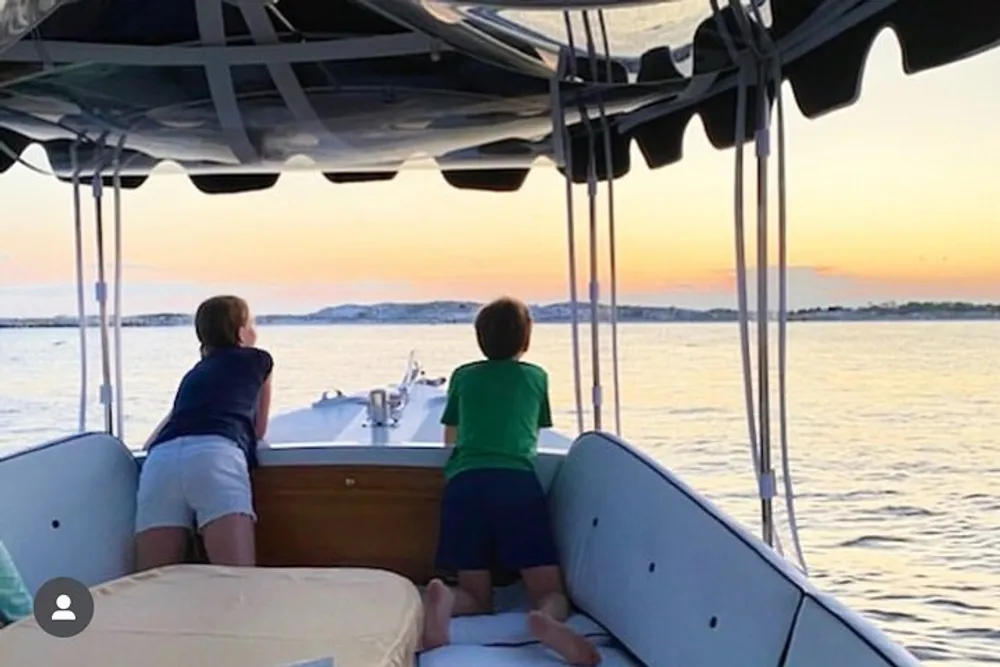 Two individuals are standing at the stern of a boat enjoying a serene sunset over the water