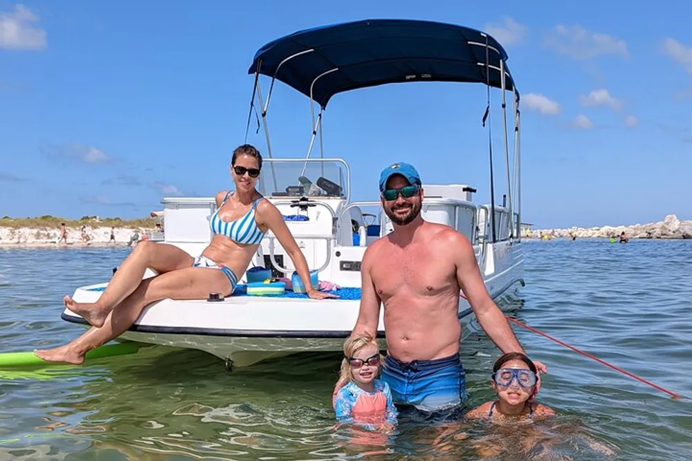 A family is enjoying a sunny day out on a boat and in the water with two children wearing snorkeling masks and a woman lounging on the boat in a bikini while a man stands in the water beside them all smiling
