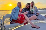 Three women are sharing a joyful moment on a boat with a beautiful sunset in the background.