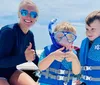 Two people wearing snorkeling gear give a thumbs-up in clear shallow water with boats and a beach in the background