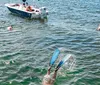Two people wearing snorkeling gear give a thumbs-up in clear shallow water with boats and a beach in the background