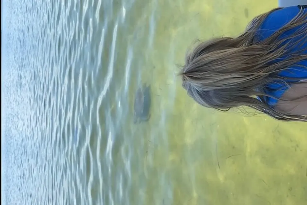 A person with long hair is peering into clear shallow water where a fish is visible