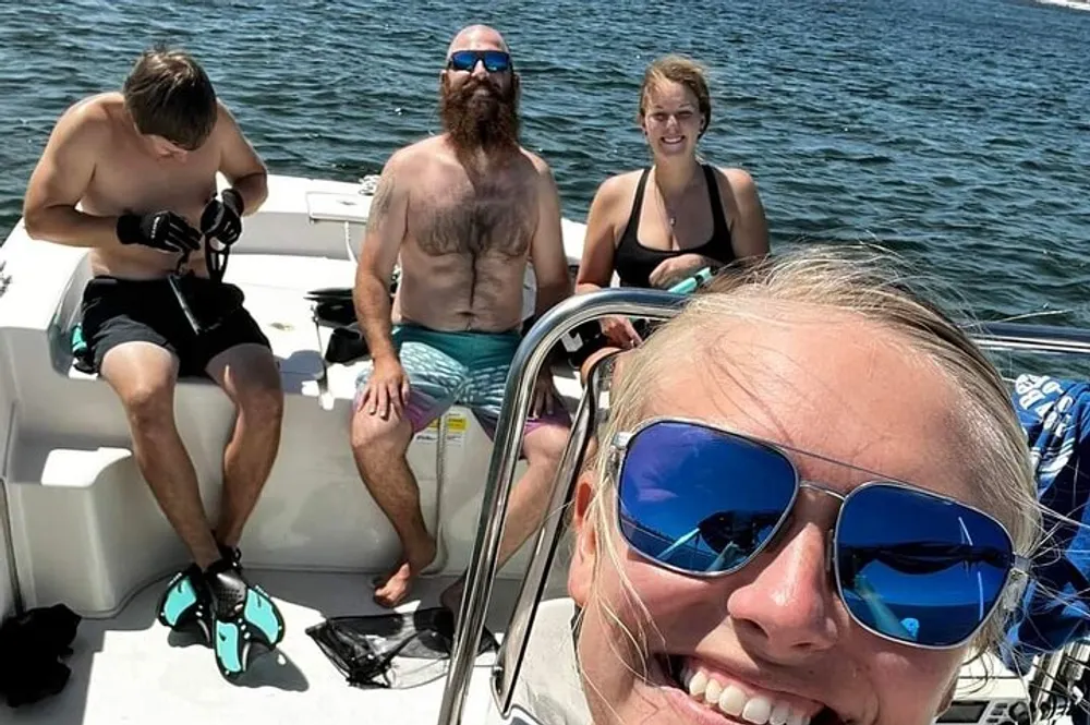 A smiling person takes a selfie on a boat with three others in the background two of whom are preparing diving gear on a sunny day