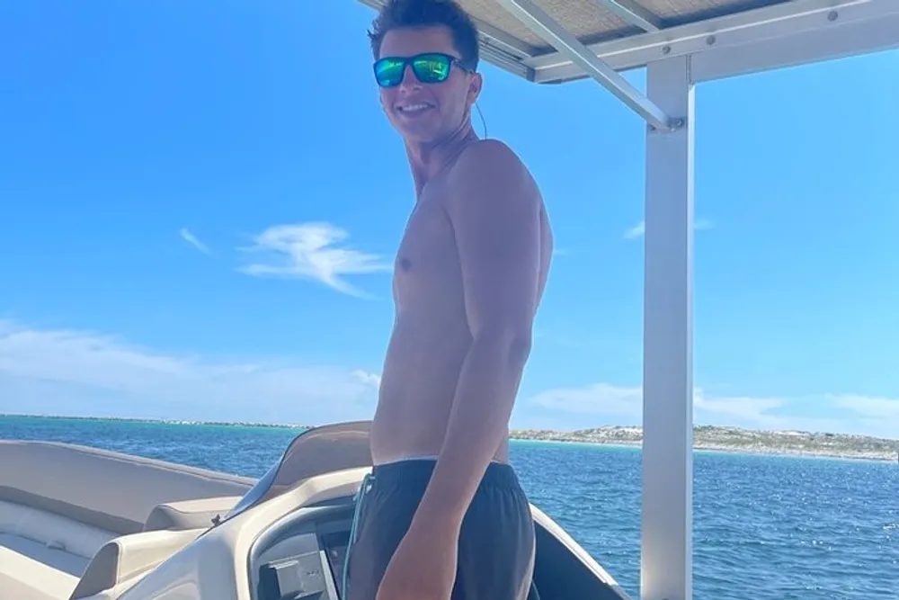 A shirtless person wearing sunglasses is smiling on a boat with clear skies and coastal scenery in the background