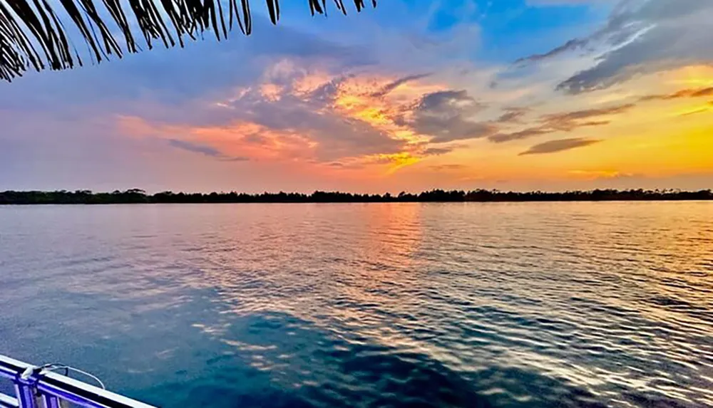 A tranquil scene captures the vibrant hues of a sunset sky reflecting off the calm waters viewed from a waterfront with a glimpse of a railing in the foreground