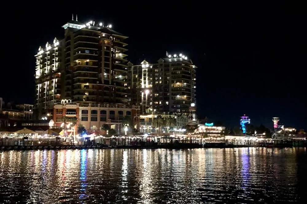 A nighttime cityscape is illuminated with the reflection of lights on the waters surface featuring high-rise buildings adjacent to a busy waterfront promenade