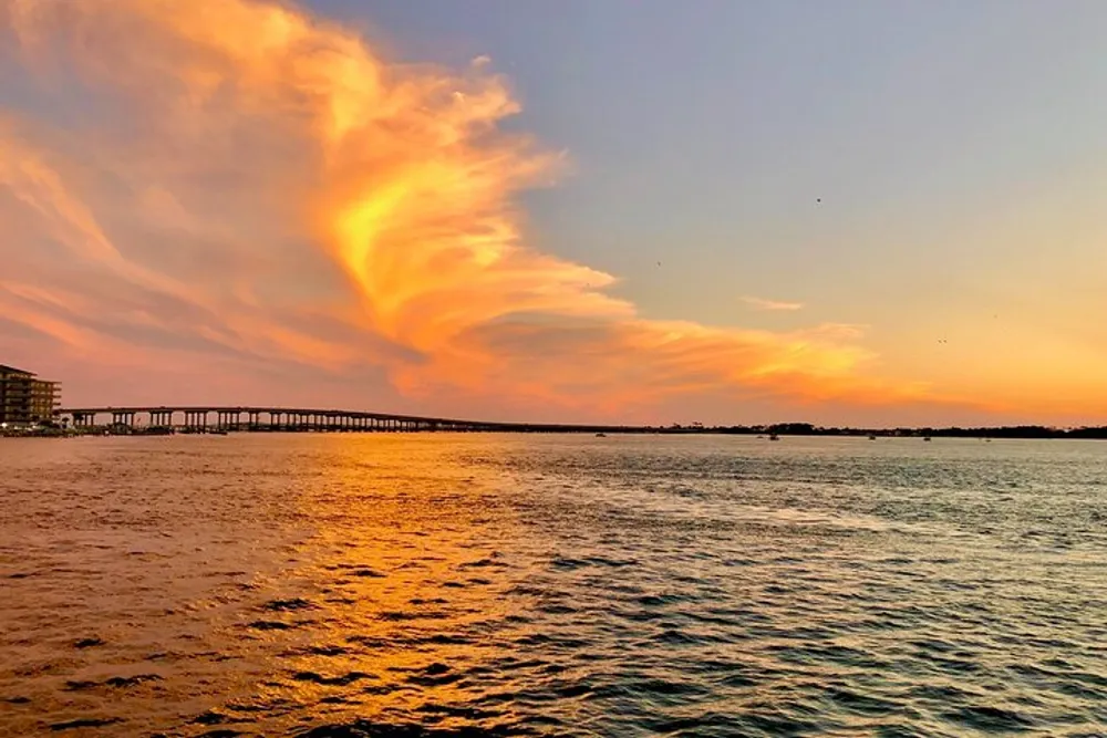 The image captures a vibrant sunset over a body of water with a fiery cloud formation in the sky and a bridge spanning the background horizon
