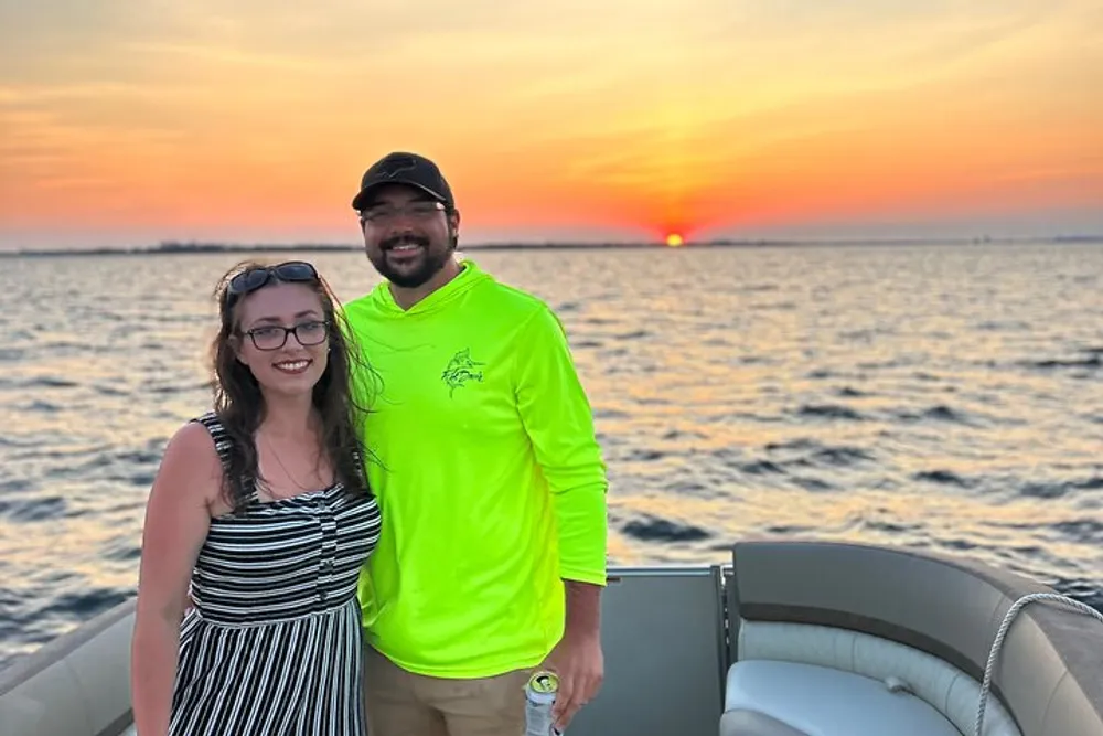 Two people are smiling for a photo aboard a boat with a beautiful sunset over the water in the background