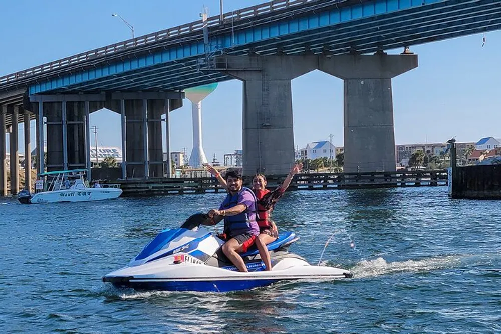 Two people are riding a jet ski on the water with a bridge and several buildings in the background