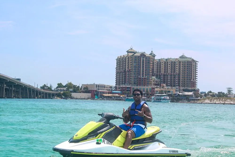A person is riding a yellow and white jet ski on turquoise water with a large building and a bridge in the background