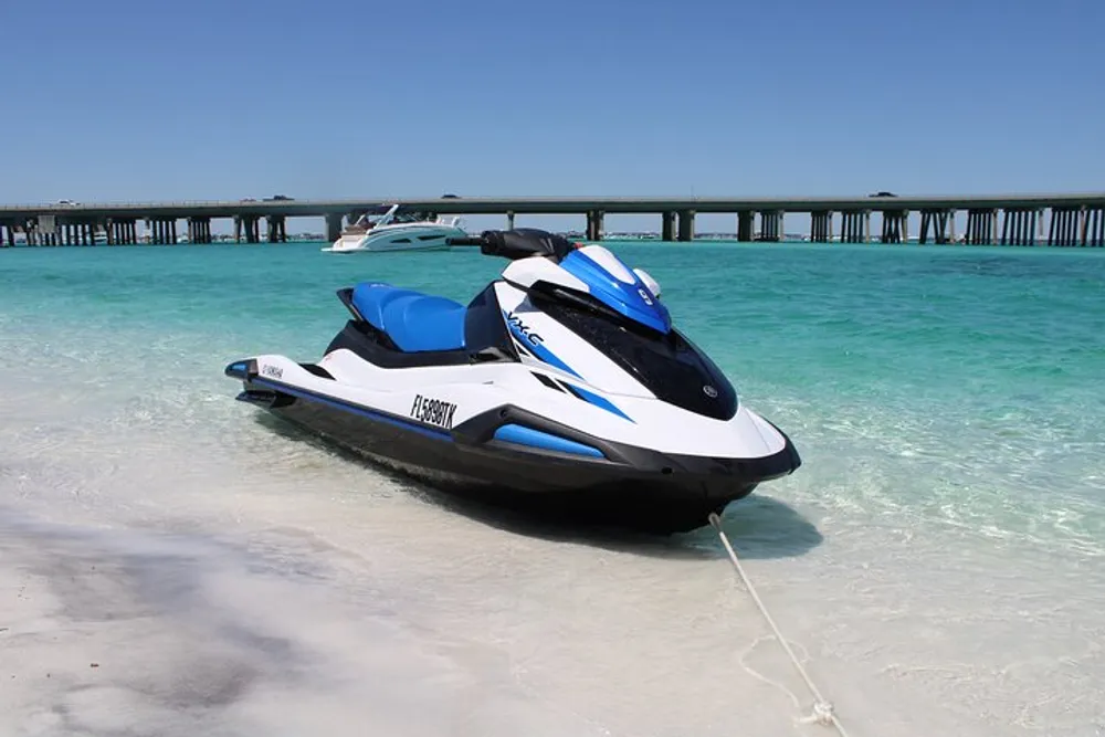 A jet ski is beached on clear shallow waters with a bridge and another vessel in the background
