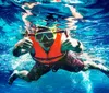 A person is submerged in clear blue water giving two thumbs up while wearing snorkeling gear and an orange life vest