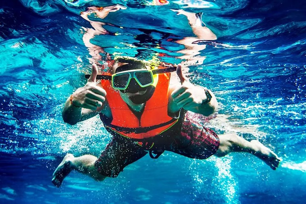 A person is submerged in clear blue water giving two thumbs up while wearing snorkeling gear and an orange life vest