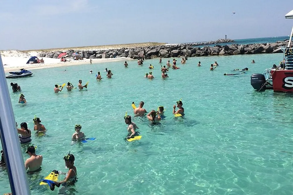 A group of people wearing snorkeling gear are wading in clear shallow waters near a beach with some on flotation devices and a motorboat anchored nearby