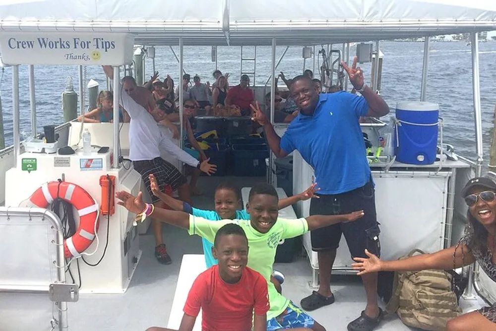 A group of people including children and adults are smiling and posing for a photo on a boat with some raising their arms in a joyful gesture