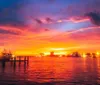 A vibrant sunset paints the sky with shades of pink orange and blue over a tranquil waterfront dotted with illuminated structures and boats