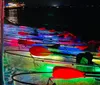 The image shows a group of illuminated transparent kayaks at nighttime glowing with a vibrant array of colors by the waters edge