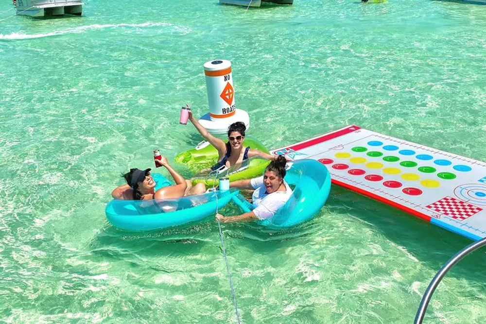 Three people are enjoying themselves on inflatable rafts one shaped like a game board and the other a floating cooler in clear turquoise waters with a No Boats buoy nearby
