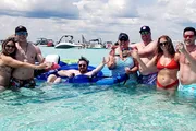 A group of people is happily posing in clear water with drinks, some on inflatable tubes, with boats visible in the background, suggesting a leisurely day at the beach or a sandbar party.