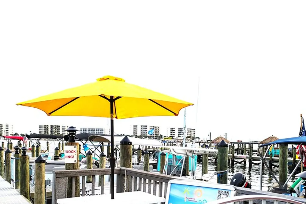 A vivid yellow parasol stands on a waterfront deck with boats and buildings visible in the background