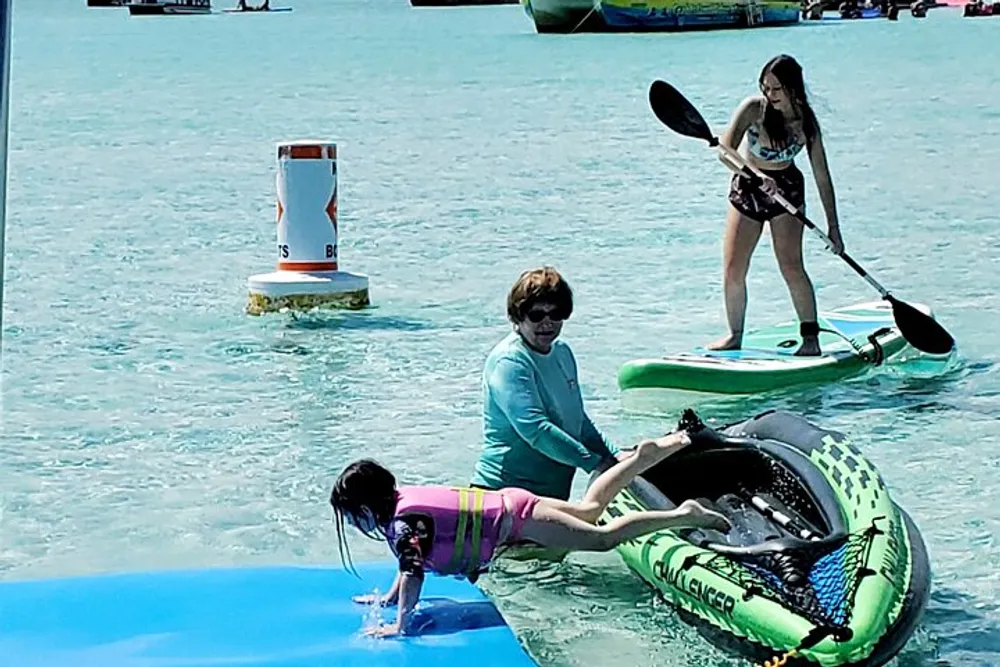 Three people are enjoying water activities with one on a paddleboard another climbing onto a floating raft and a third one seated on another raft in a bright sunny setting